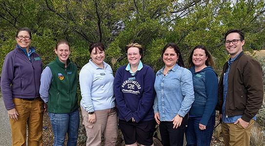 A group of seven Day Camp Teachers standing in front of green shrubbery. The sun is shining and they are all smiling broadly.